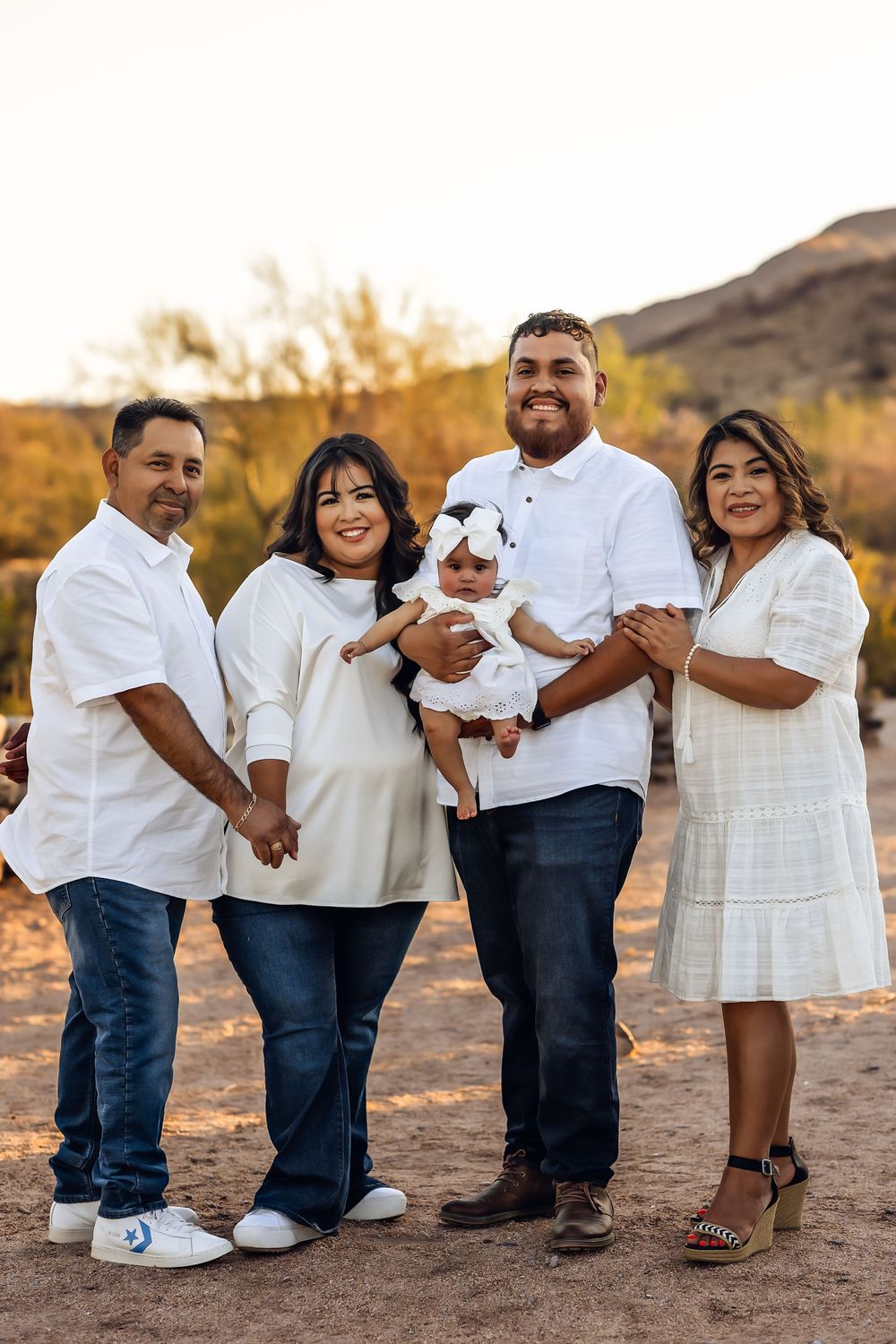 Family wearing white and cream colored outfits poses together outdoors in desert setting during sunset.