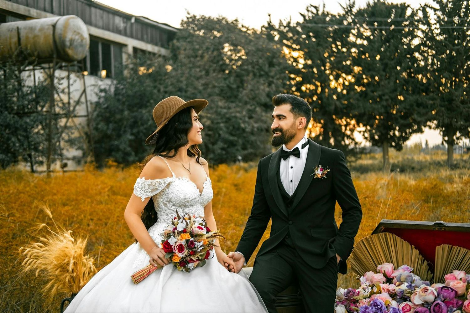 A couple in wedding attire stands together in a golden field at sunset with decorative floral arrangements nearby.