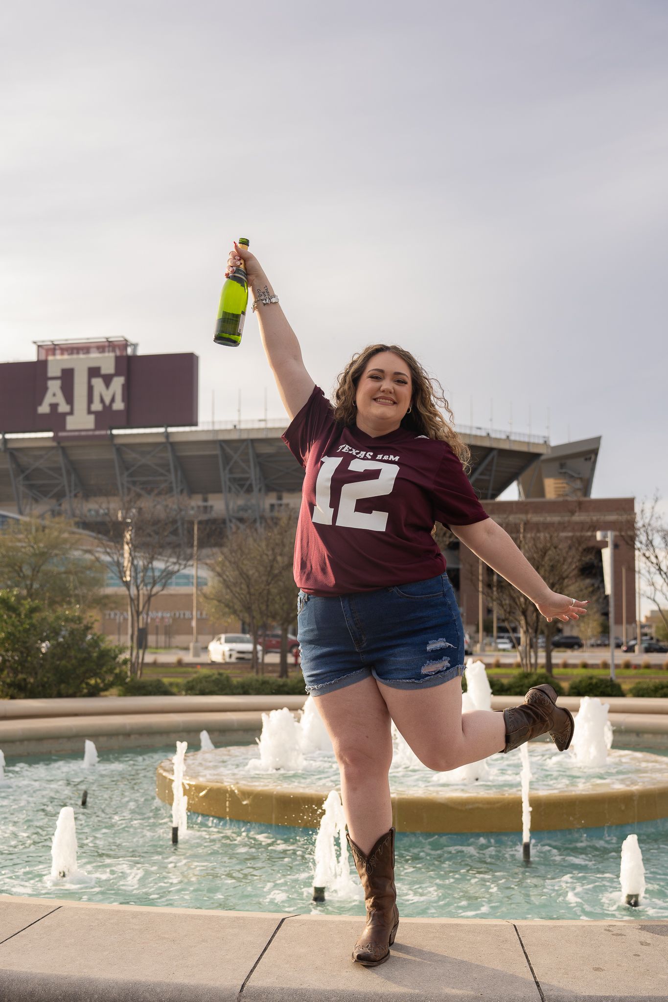 A Texas A&M graduate in a black dress and maroon stole poses by a fountain on campus during graduation season.