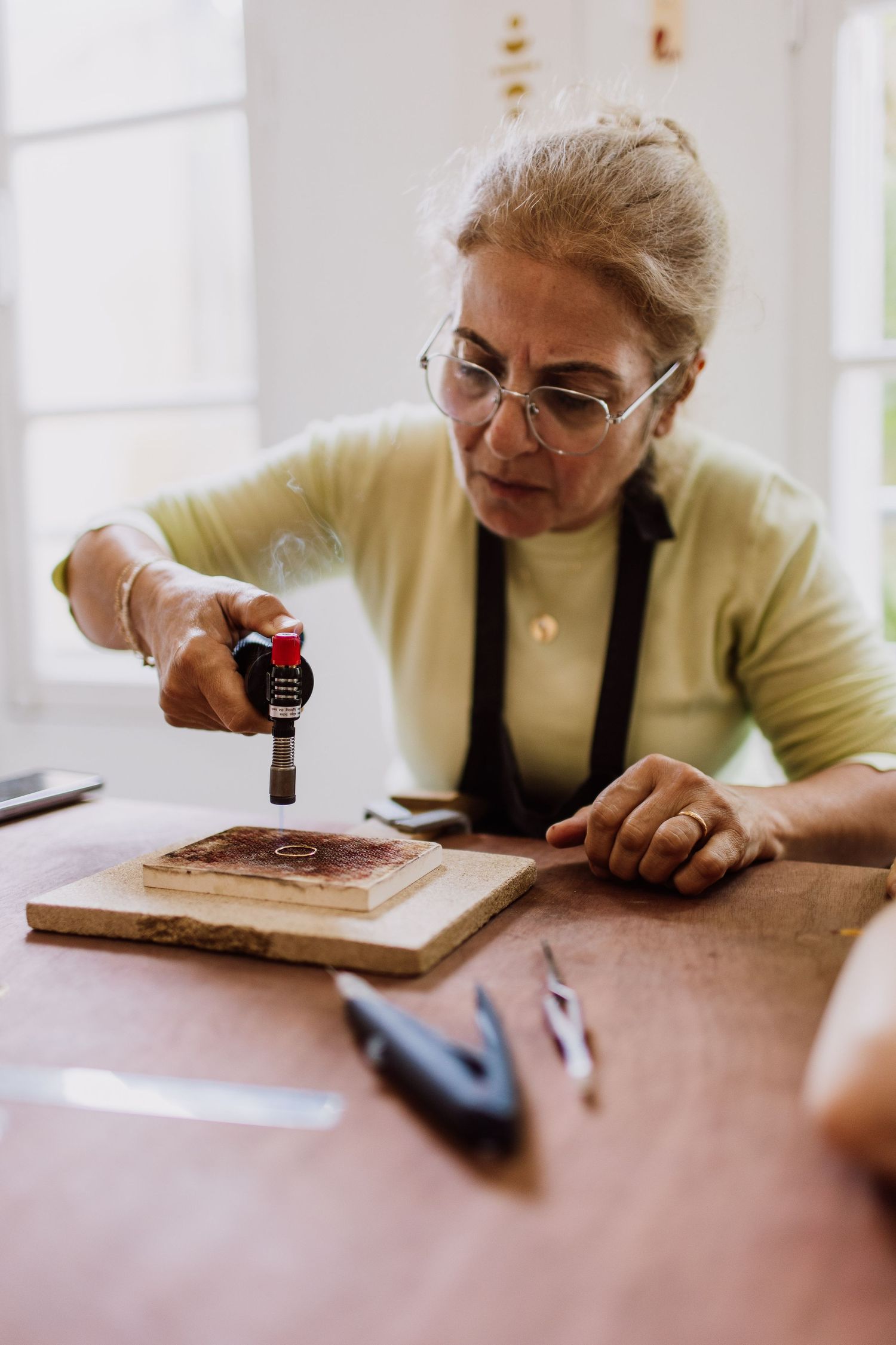 Artisan wearing glasses and yellow sweater uses tools on a wooden workbench during jewelry making.