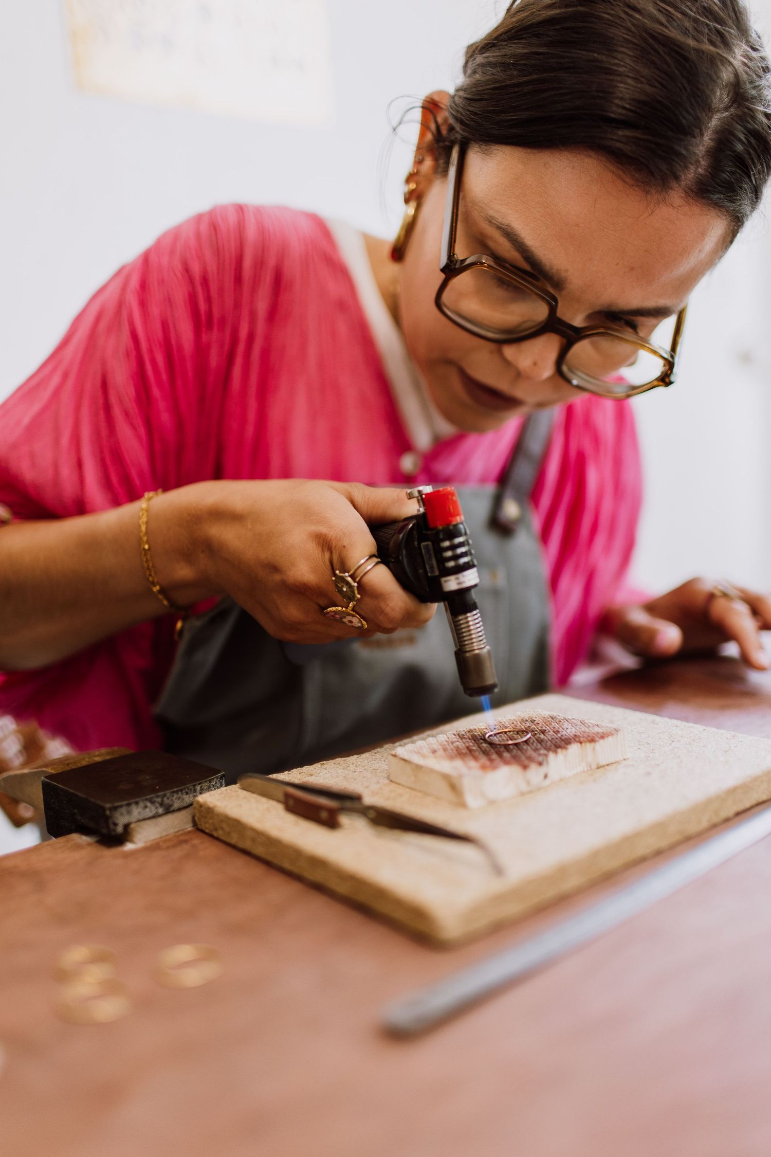 Jewelry maker in pink shirt and apron uses precision tools on a workbench.