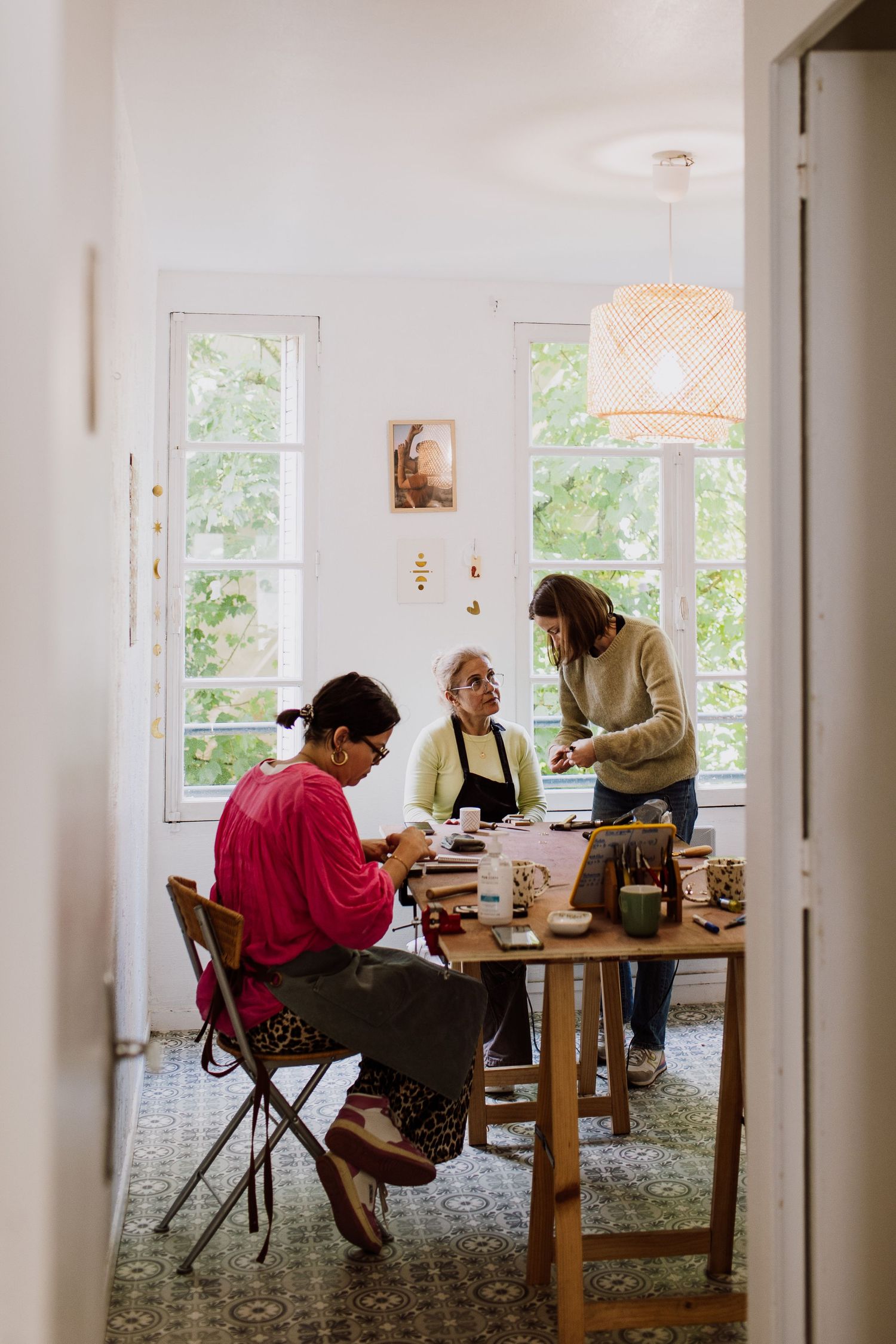 Bright sunlit crafting workshop with people working at a wooden table near windows.