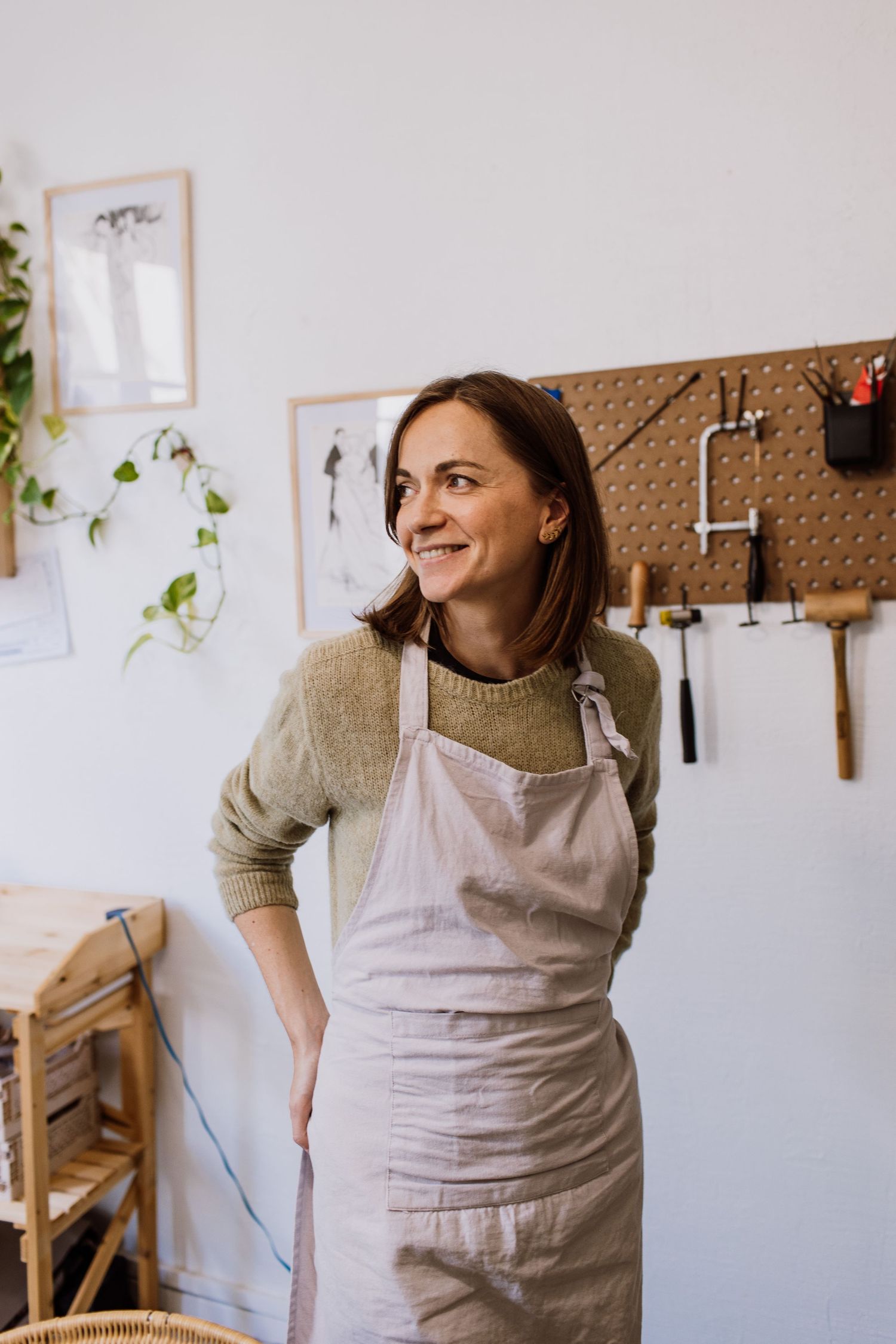 Artist in workshop wearing beige sweater and apron smiling against white wall with tools.