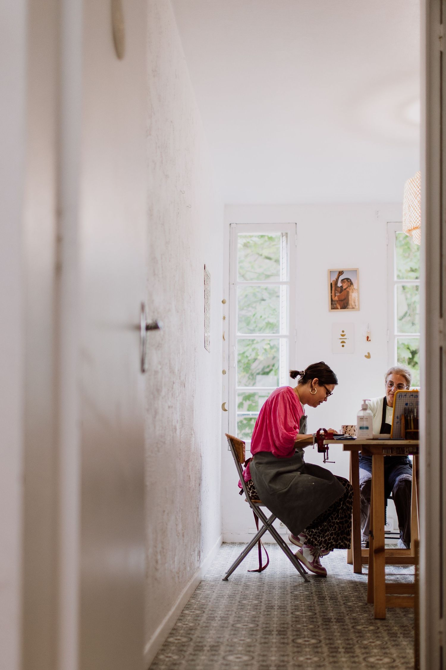 Sunlit workspace with people sitting at wooden table near white walls and windows.