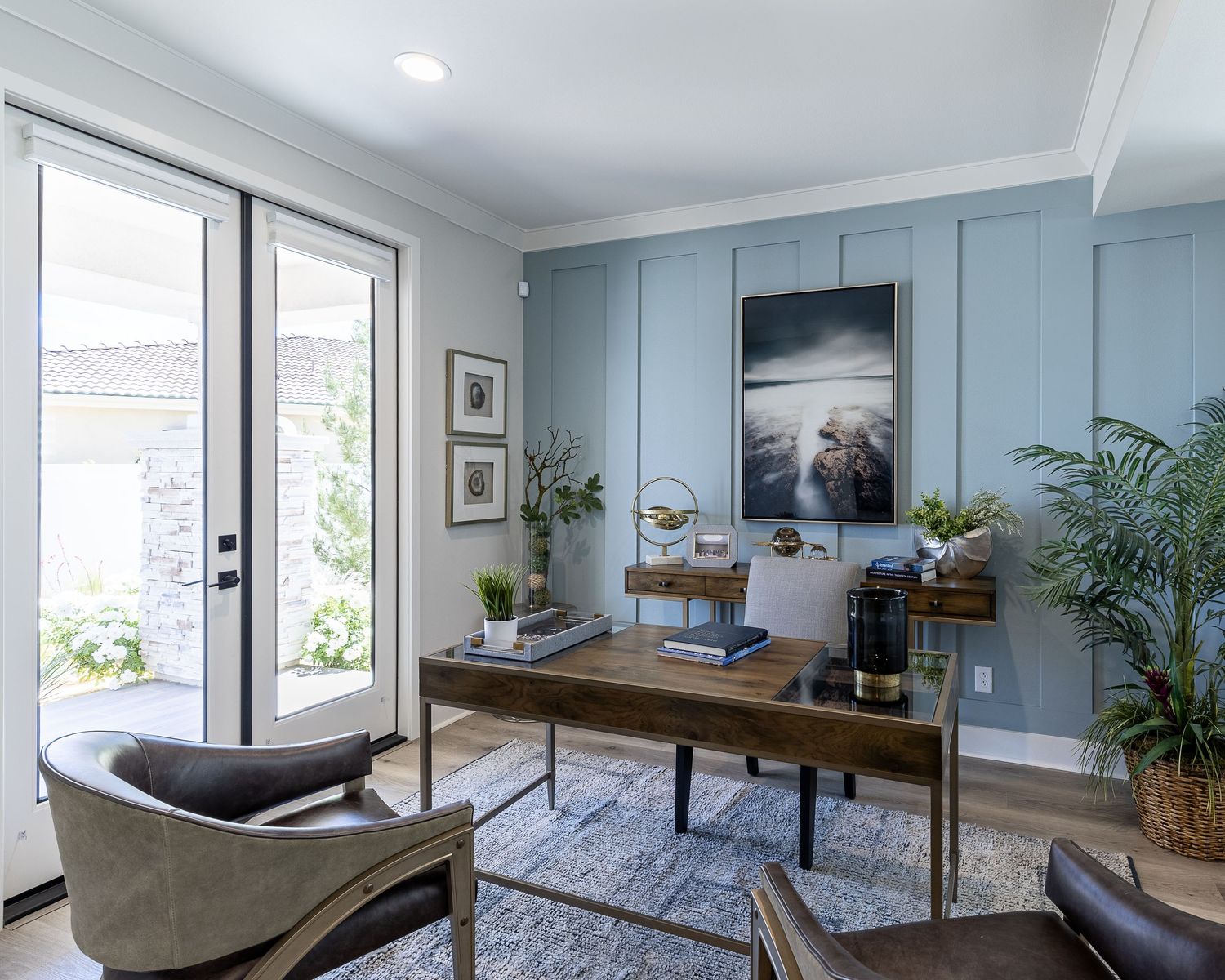 Modern home office with light blue walls, wood desk, glass doors, potted plants and a gray patterned area rug.