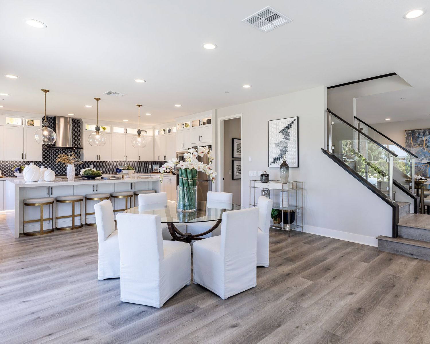 Modern open concept living space with white kitchen, dining area and staircase featuring warm wood flooring and recessed lighting.