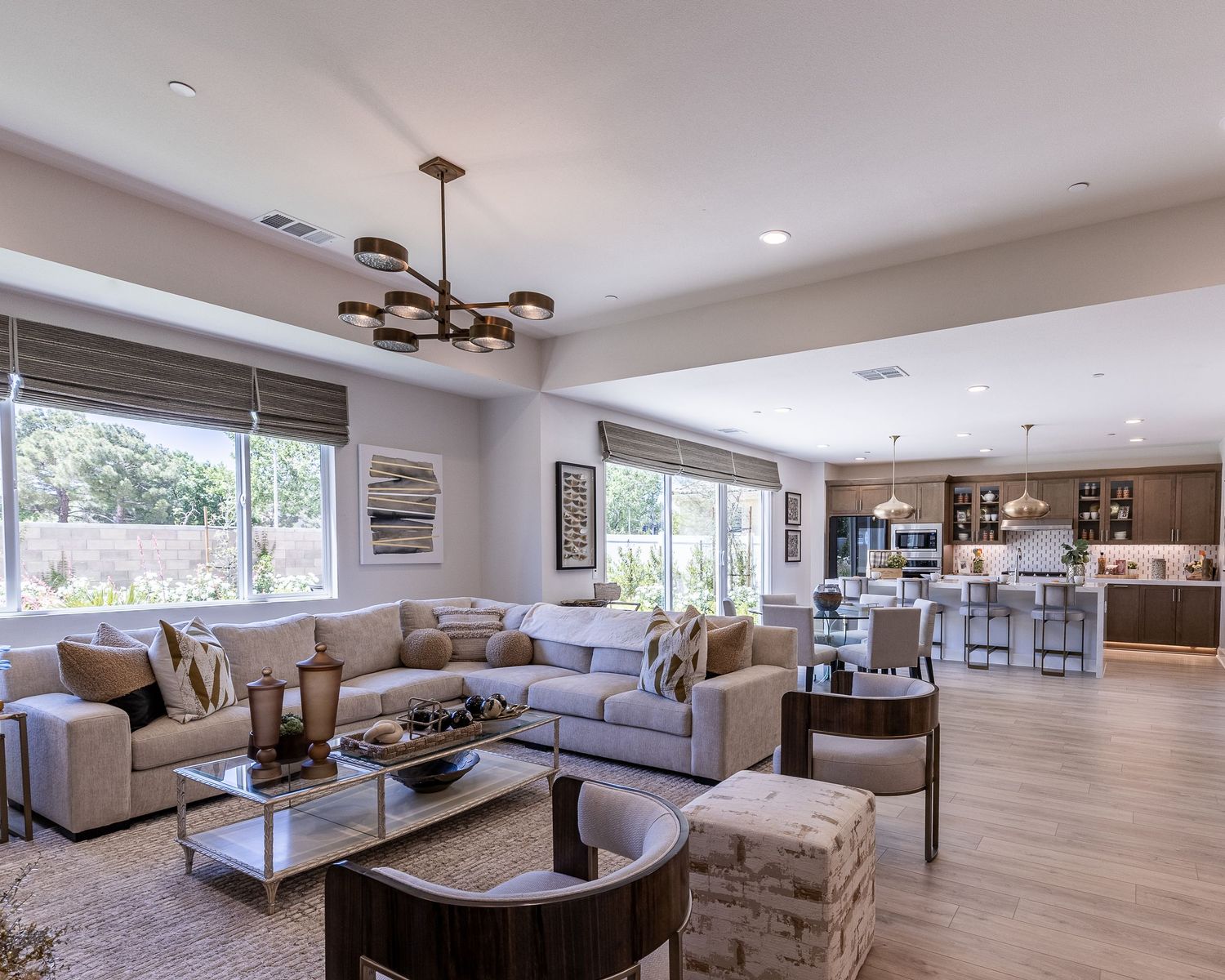 Modern open concept living room with beige sectional sofa, contemporary lighting, and connected kitchen featuring wood cabinets.