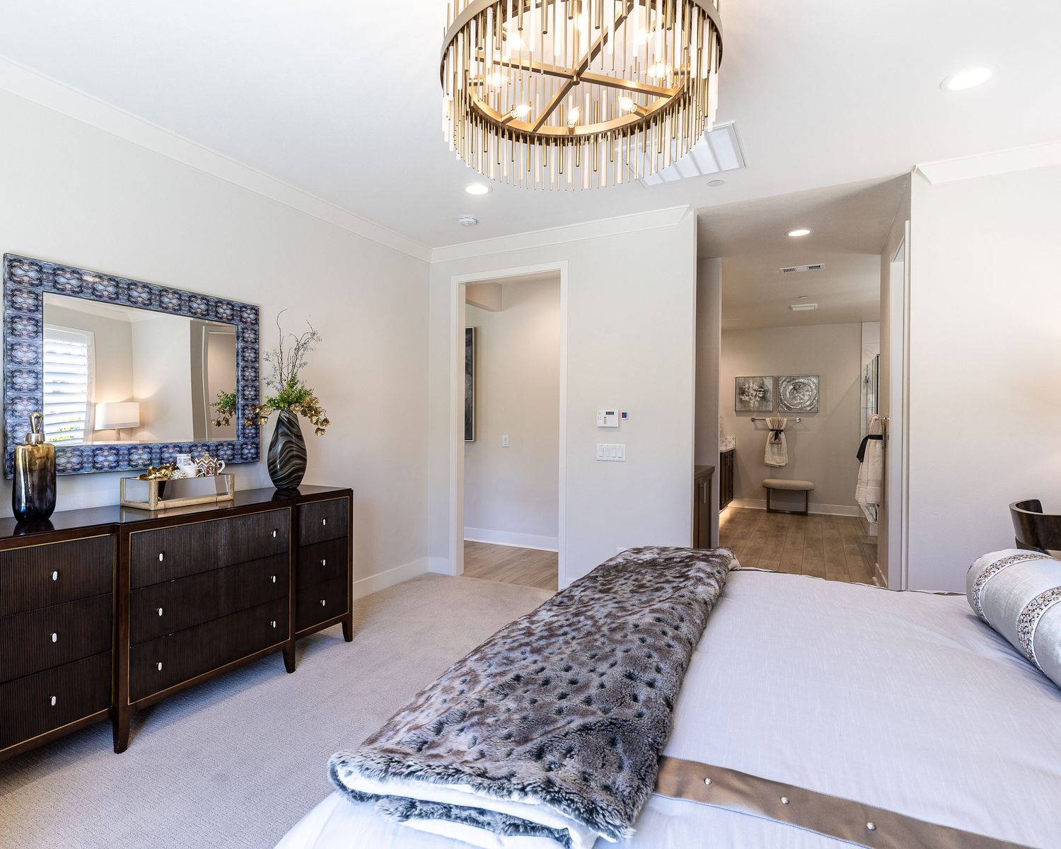 Modern master bedroom featuring dark wood dresser, circular chandelier, and plush bedding in neutral tones.