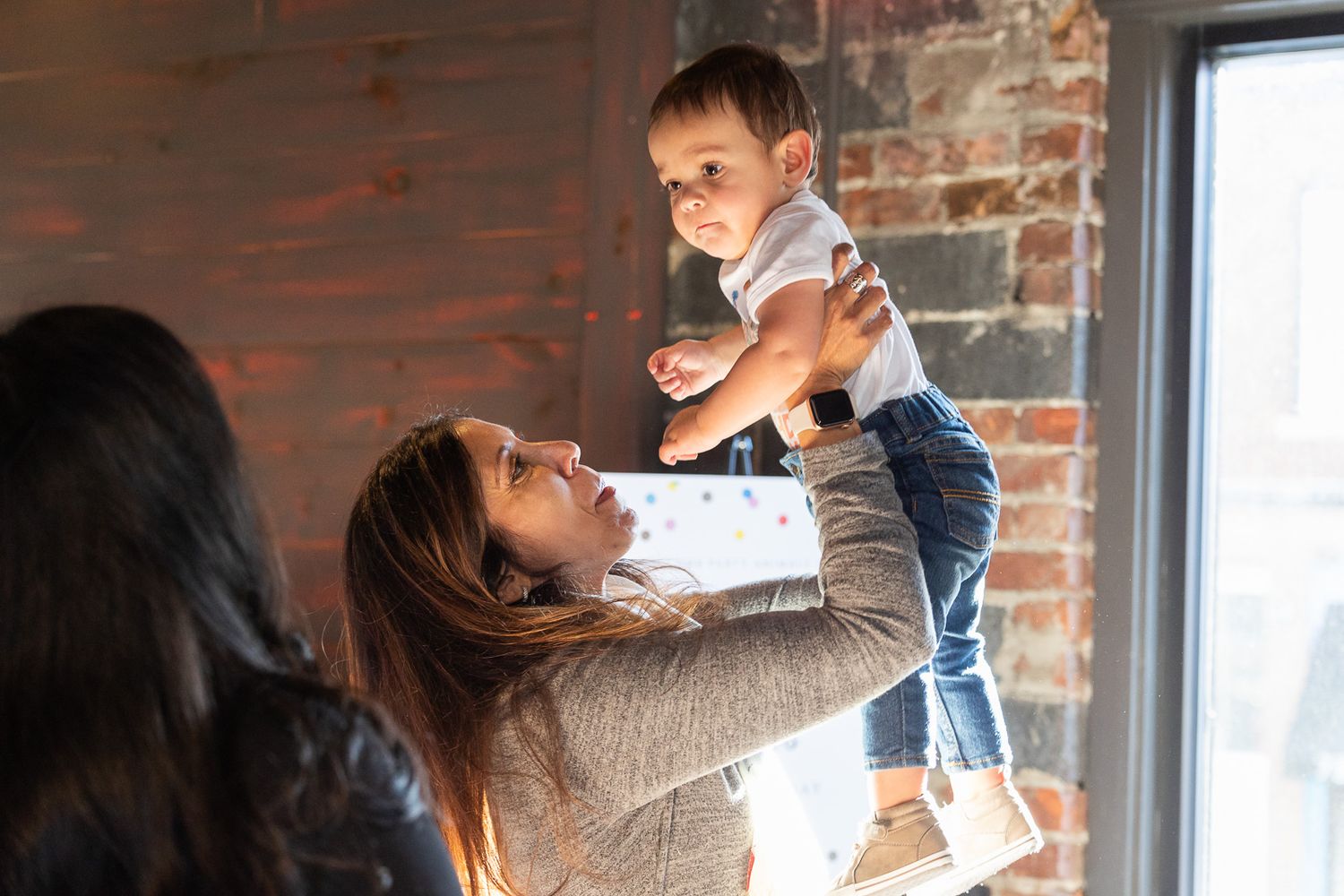 Joyful indoor gathering with lifted child against exposed brick wall and window.