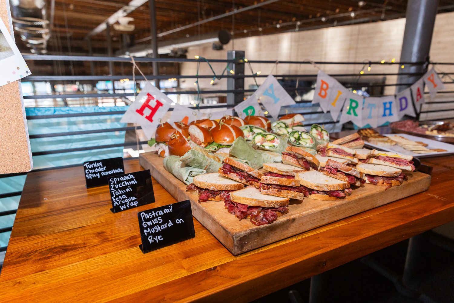Wooden board displaying assorted sandwiches with small identifying signs at a catered event.