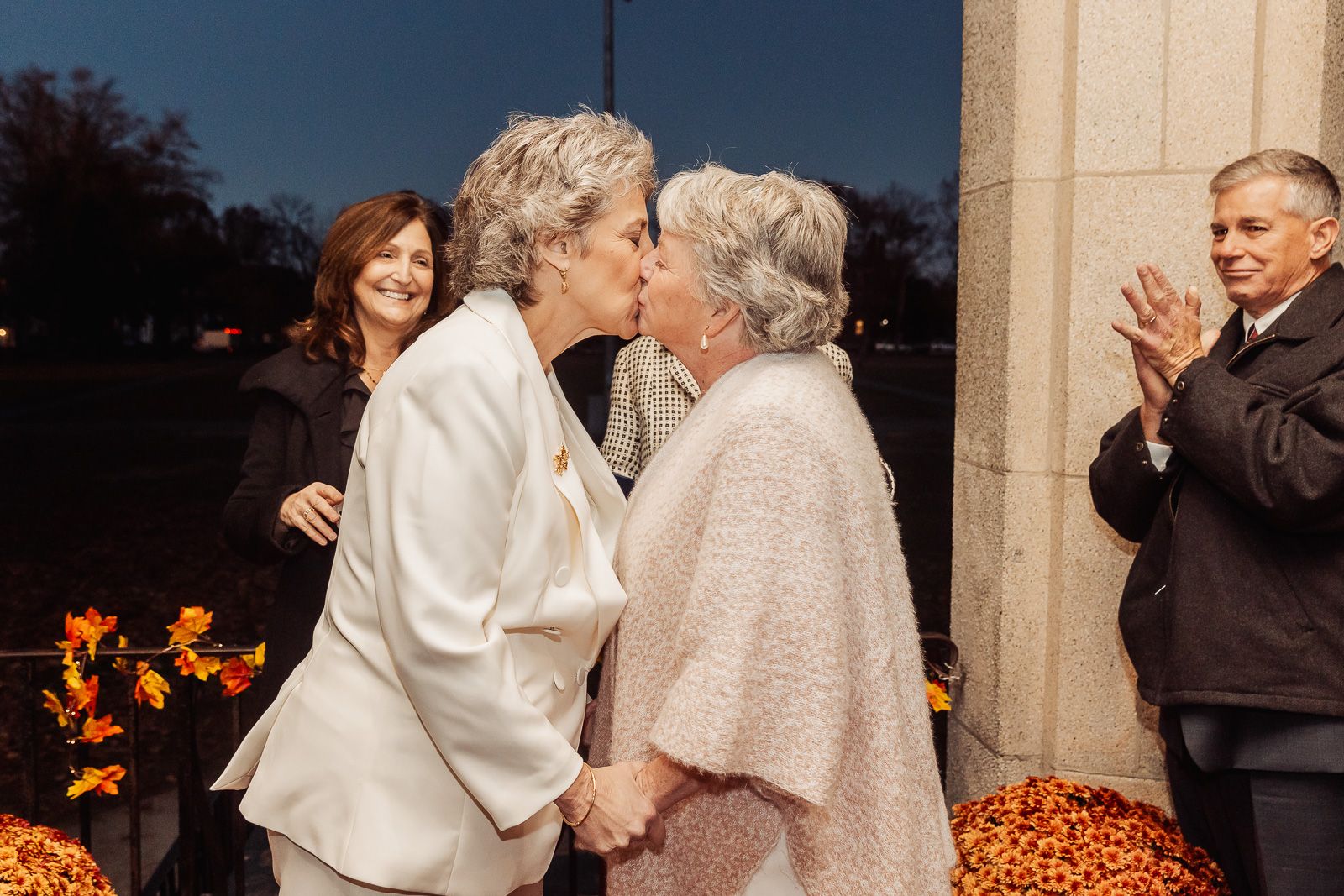 A tender wedding moment captured as a couple shares a kiss while guests applaud in the background.