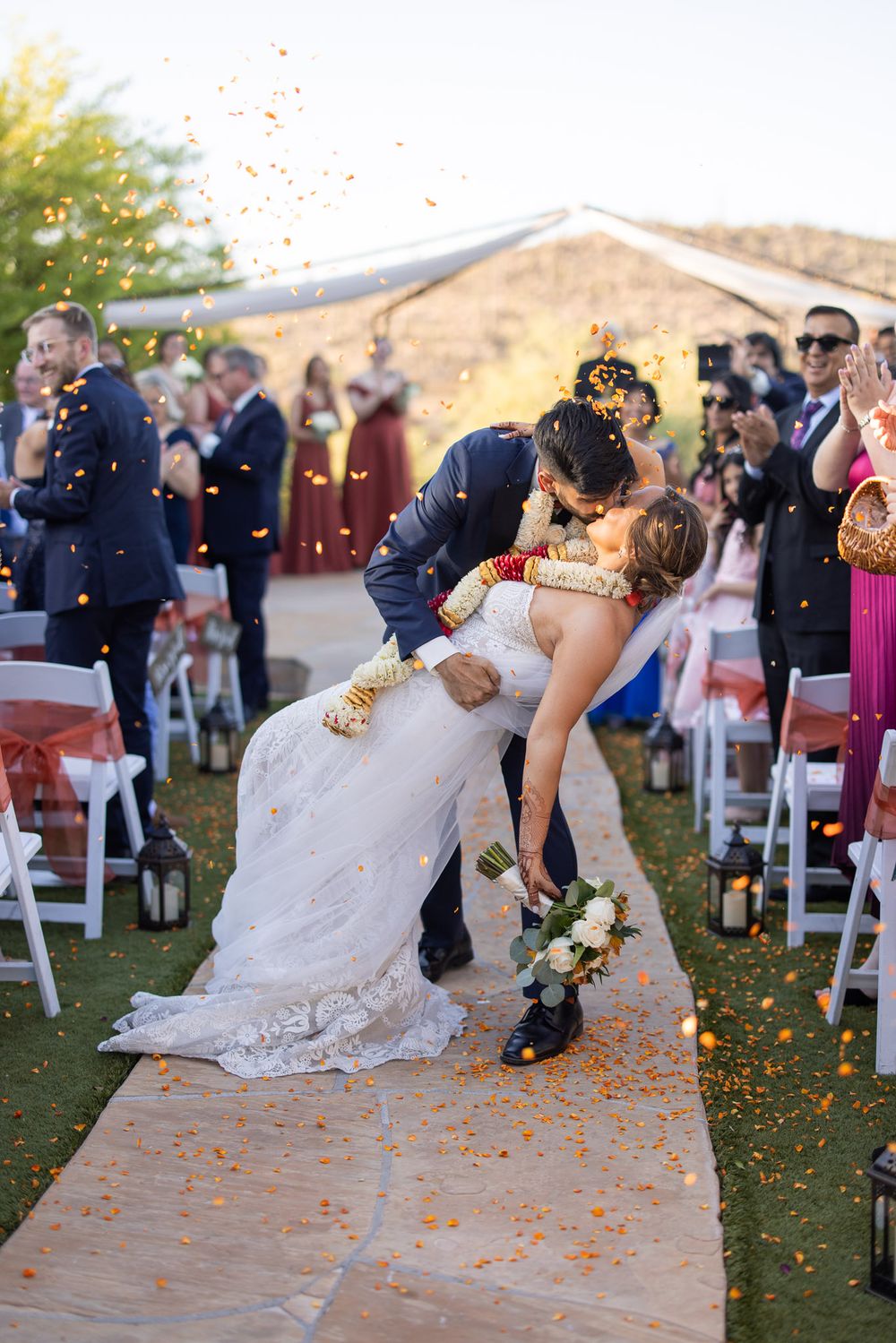 A romantic wedding kiss on an outdoor aisle runner with orange flower petals falling and guests watching in the background.