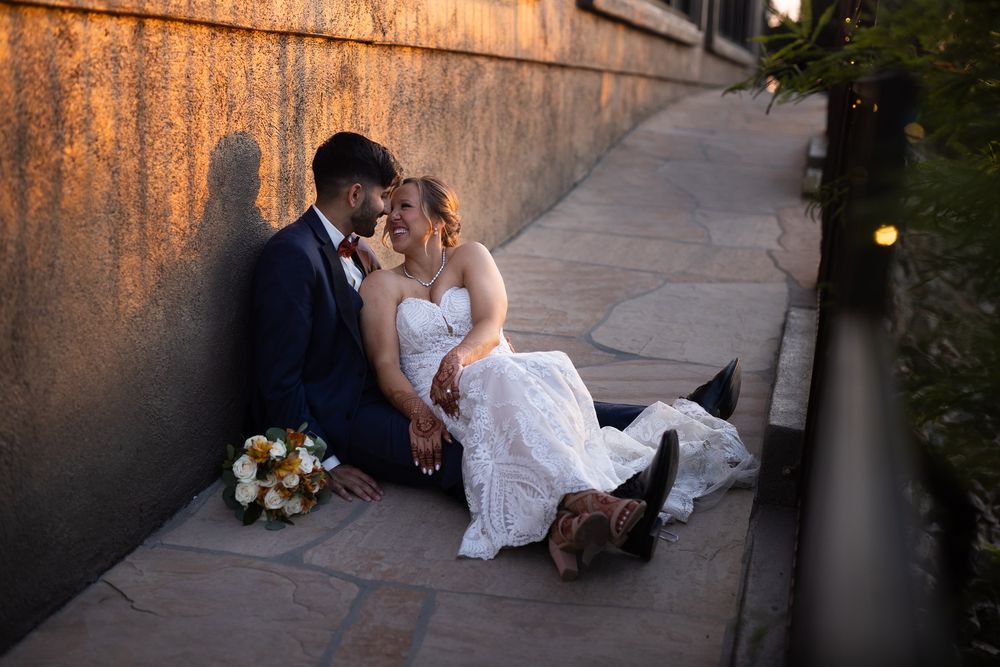 A romantic newlywed couple sits together at sunset with a bouquet of flowers beside them on stone steps.