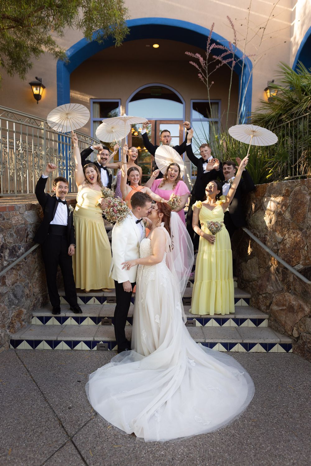 Wedding party celebrating on ornate stone steps with decorative parasols against Spanish-style archway entrance.