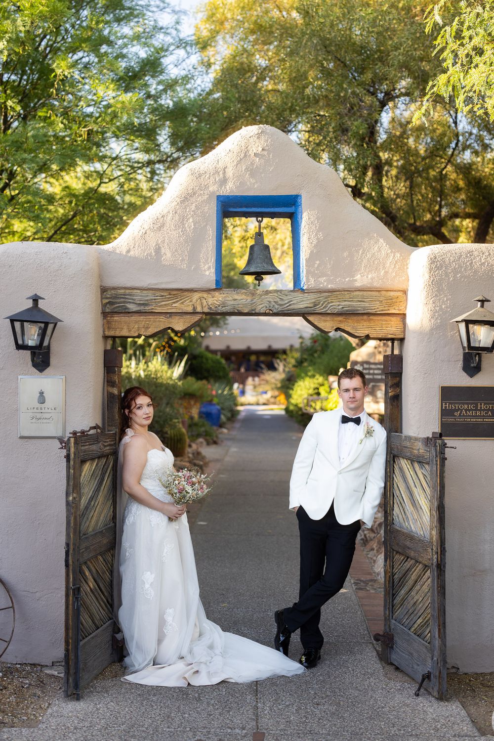 Wedding couple poses at a Spanish colonial courtyard entrance with adobe walls and lanterns at sunset.