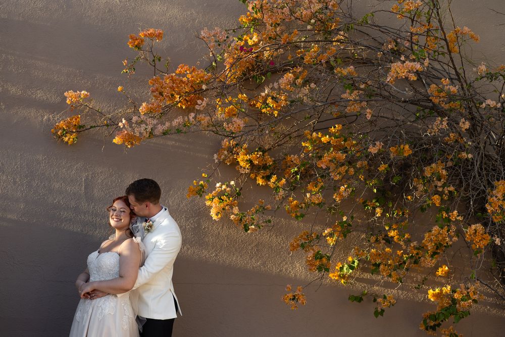 Elegant couple embraces under blooming orange bougainvillea flowers against a textured wall.