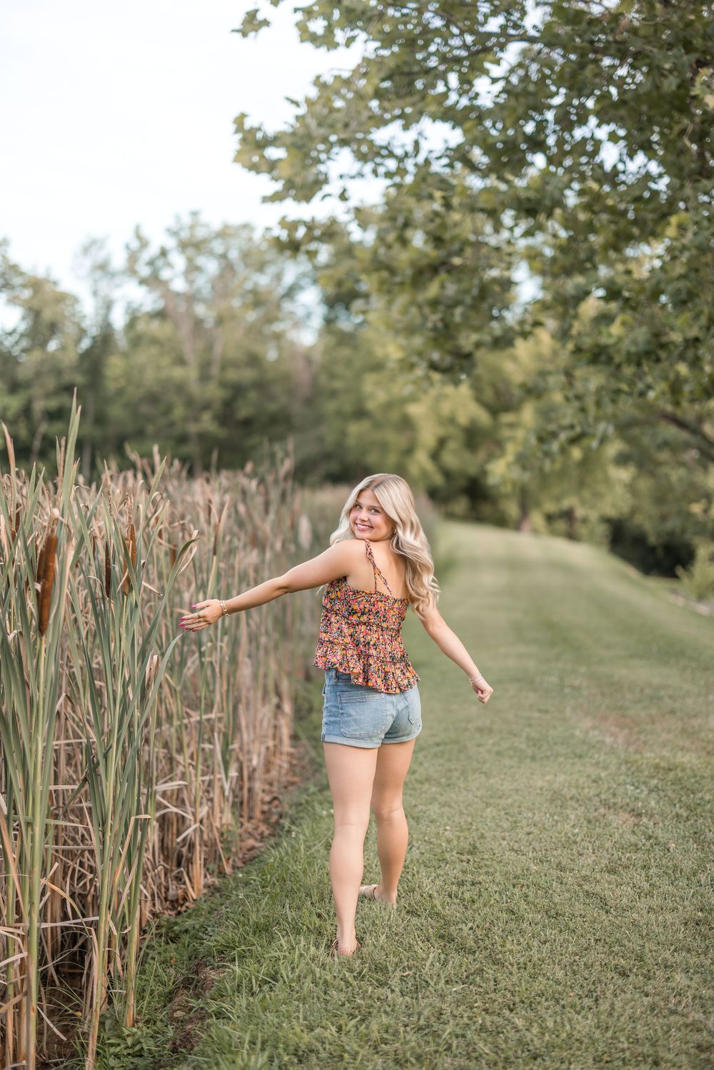 Person in denim shorts and floral top dancing barefoot along a grassy path beside tall cattails and trees.