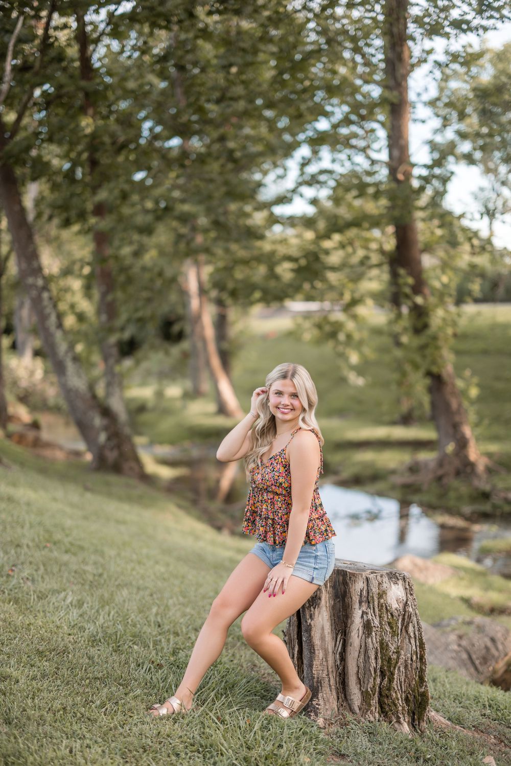 A rustic summer portrait session near a creek with a floral top and denim shorts on a tree stump.