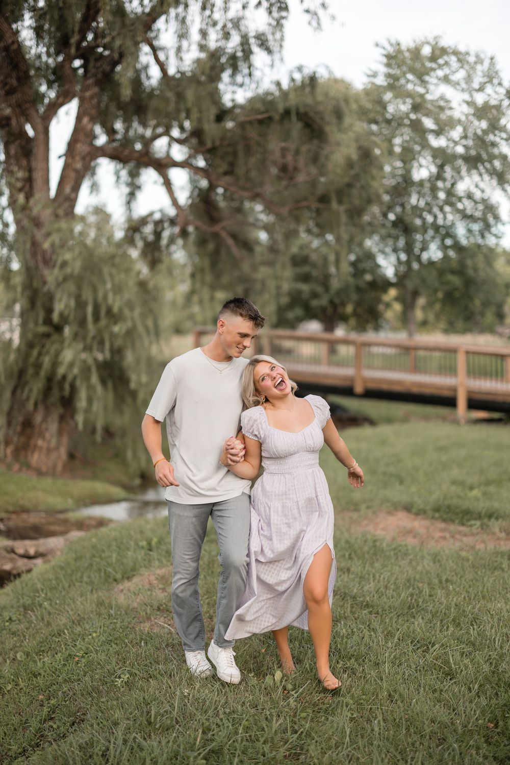 Romantic couple embracing and laughing together on grass near a wooden bridge.