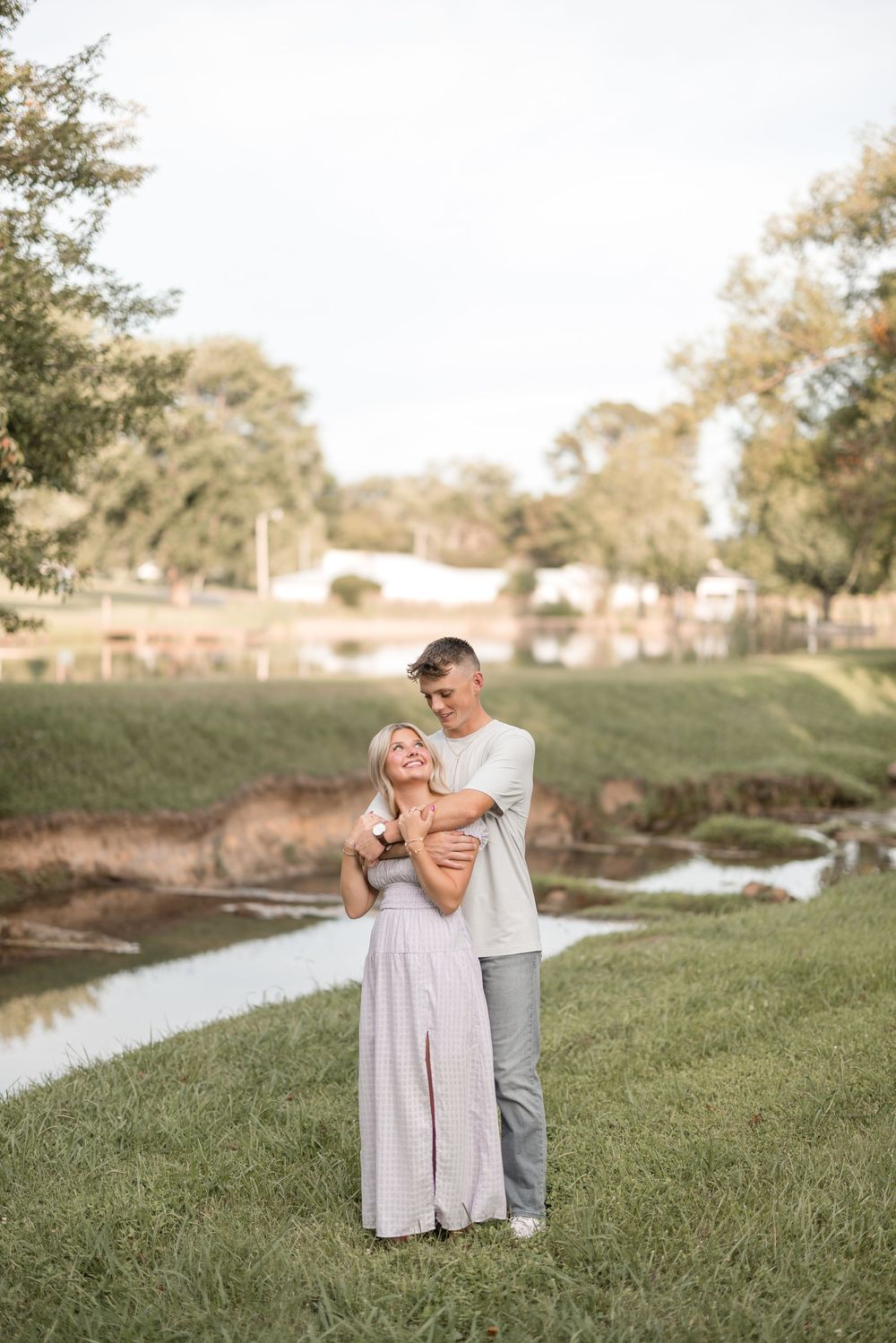 Couple in casual attire sharing an intimate moment on grassy riverbank at sunset.