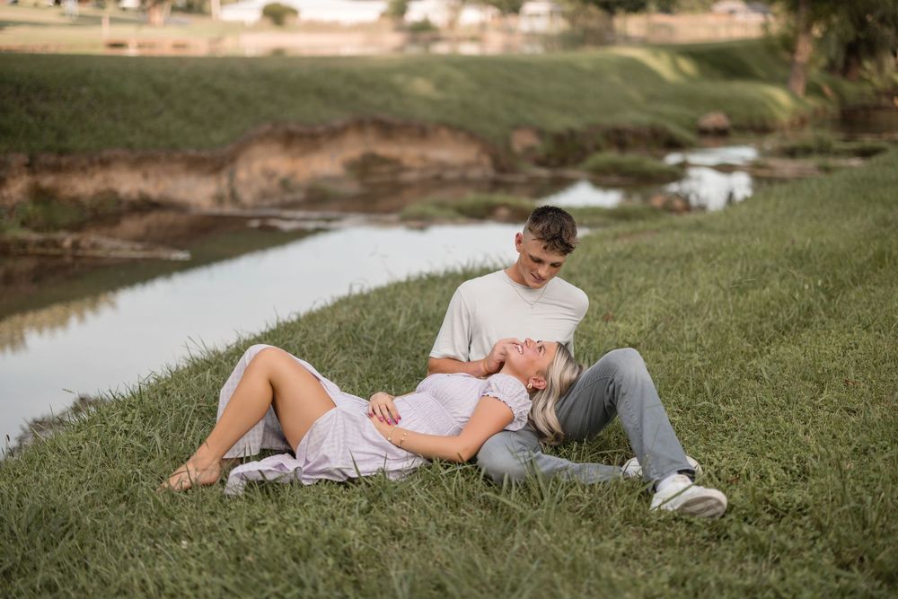 Couple relaxing on grassy riverbank during golden hour wearing casual clothing.
