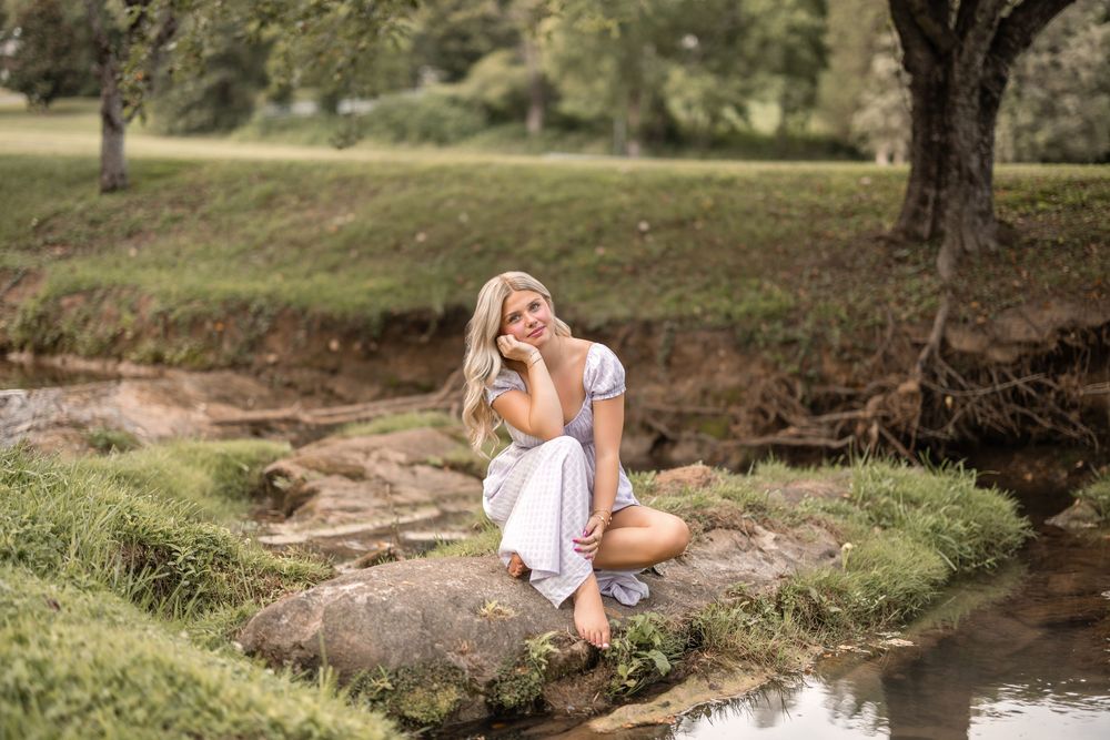 A person in a white dress sits peacefully on rocks by a tranquil stream in a sunny park setting.