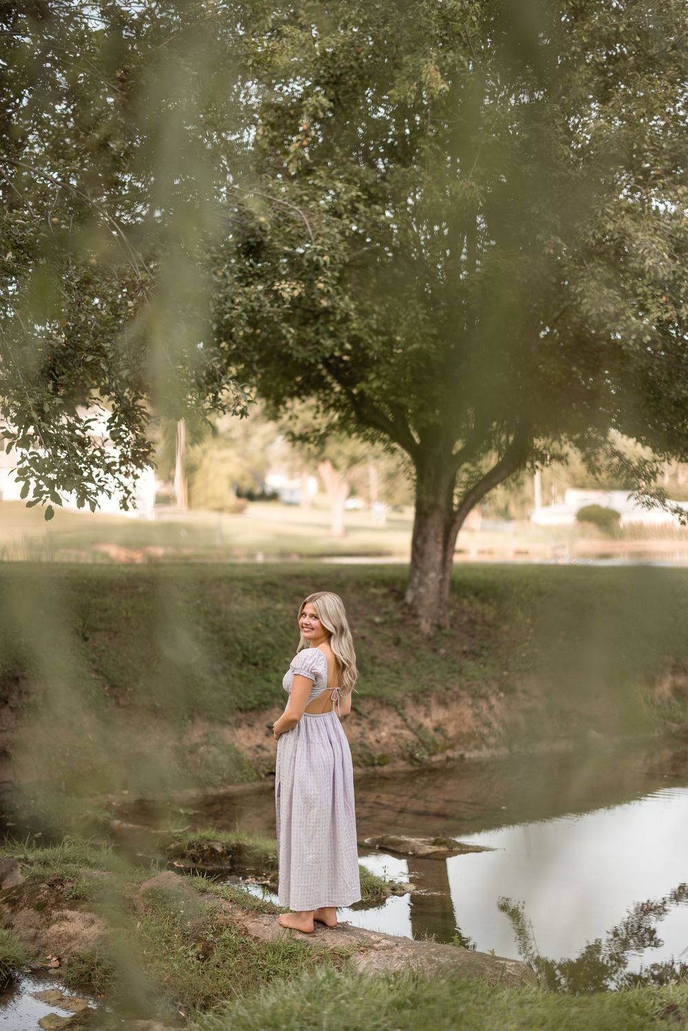 Person in a long dress stands at the edge of a tranquil pond surrounded by trees in a dreamy setting.