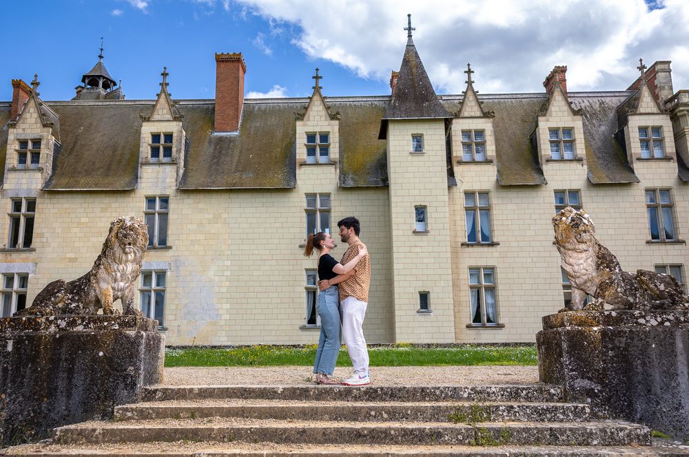 A couple stands between large stone cheetah statues on the steps of a historic French chateau with dormer windows.