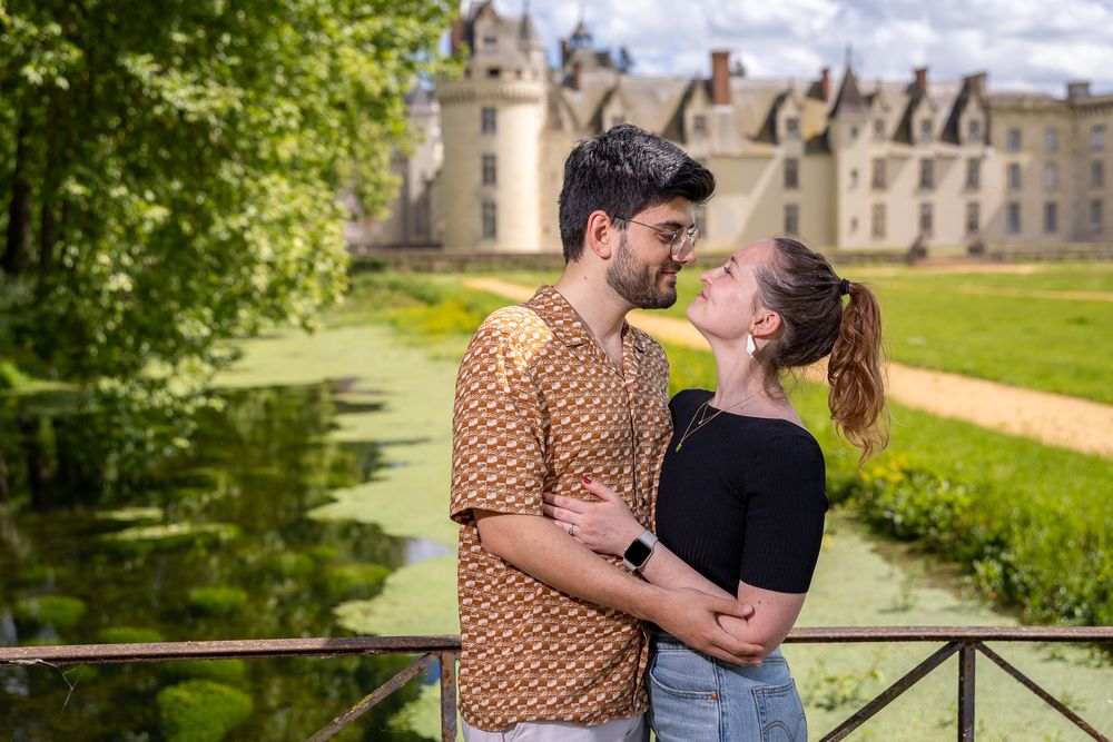 A happy couple embraces on a bridge railing with a majestic French chateau in the background on a sunny day.