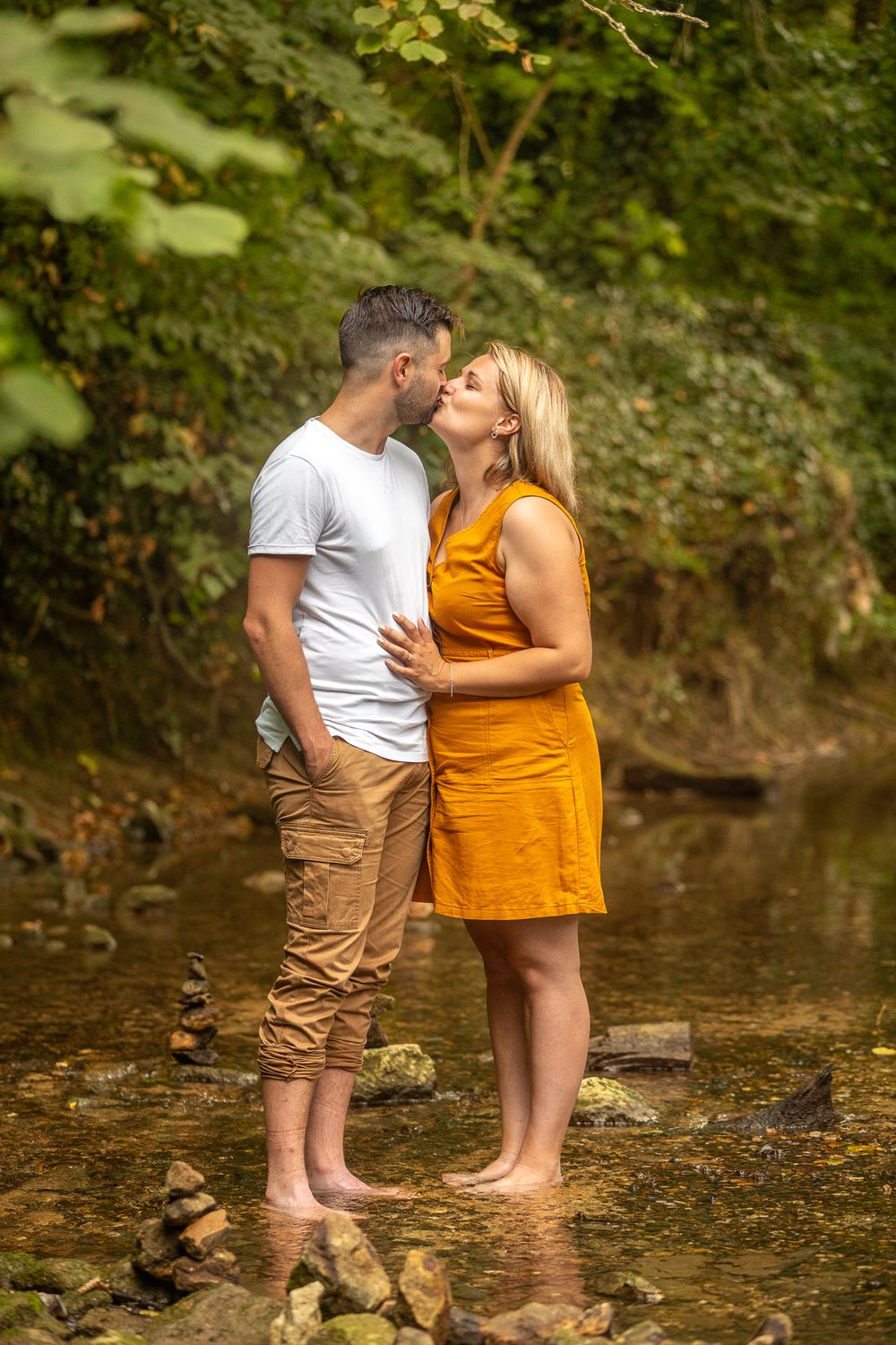 Couple standing barefoot in a shallow creek surrounded by lush greenery during a romantic outdoor photoshoot.