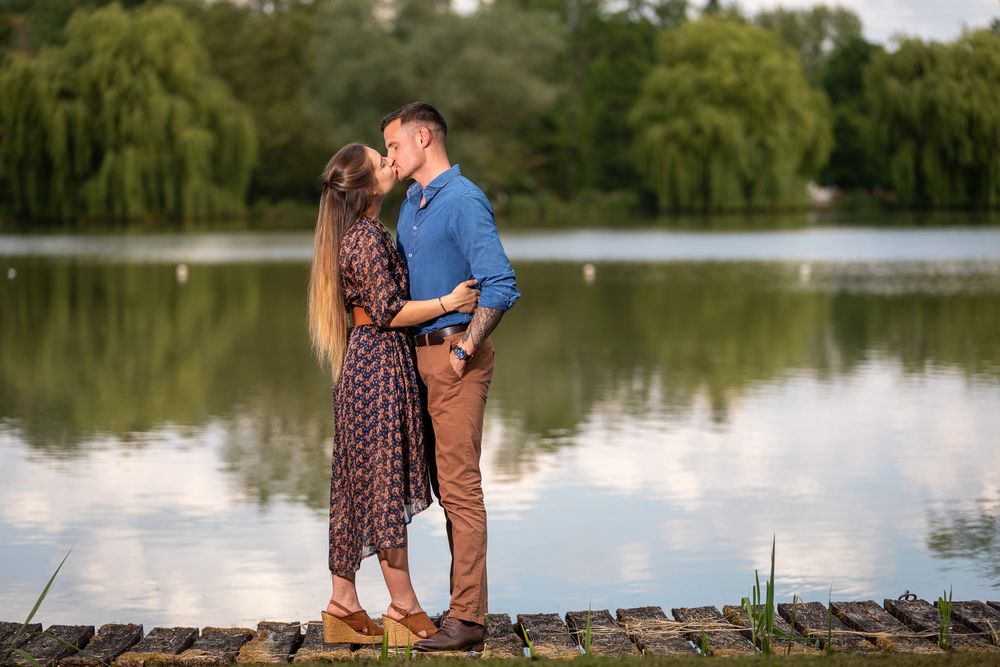 A couple embraces on a wooden dock beside a serene lake with weeping willow trees in the background during sunset.