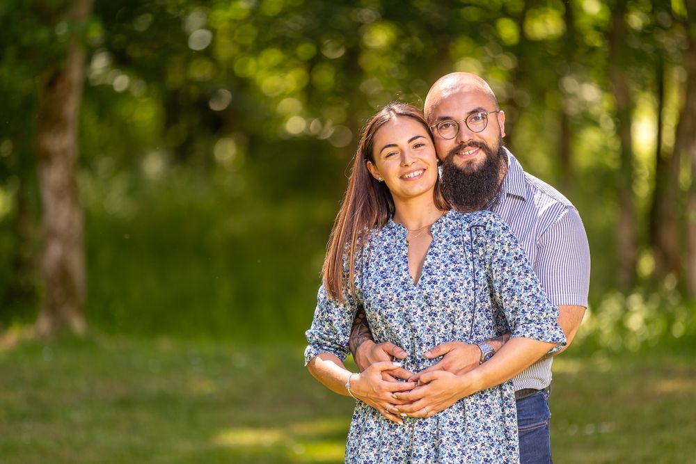 Couple in casual attire embracing and smiling during outdoor photo session in sunlit wooded area.