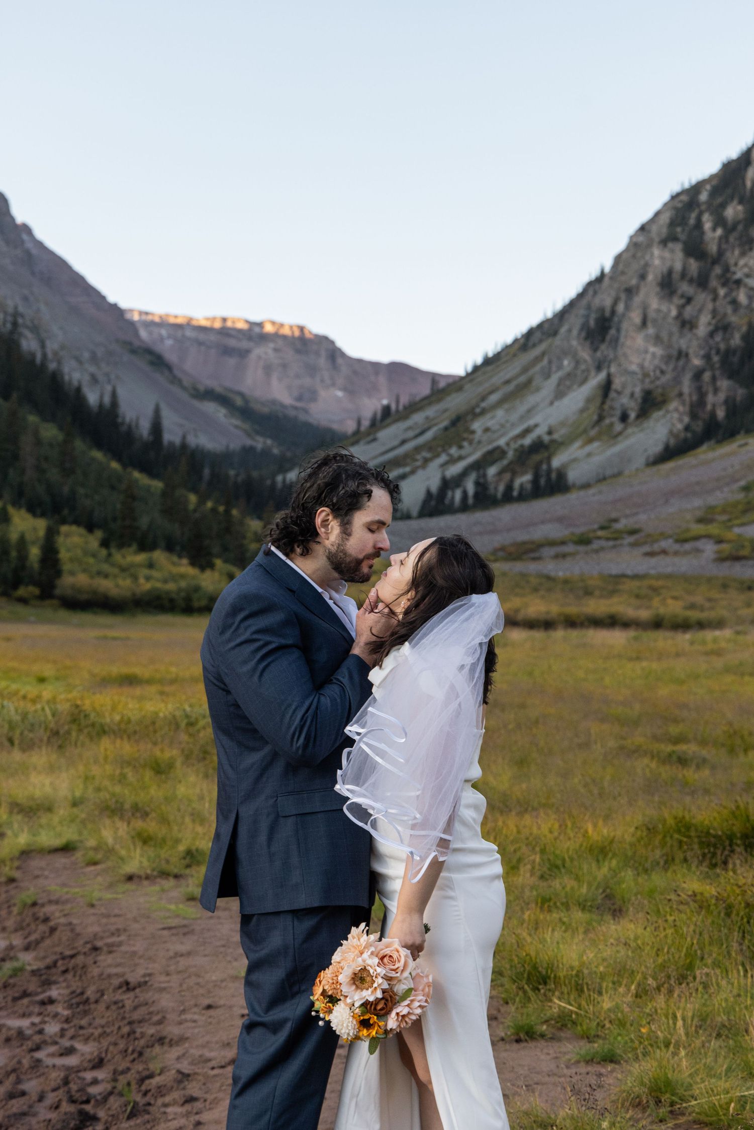 A romantic wedding photo captured in a valley with dramatic mountain peaks and golden sunlight illuminating the landscape.