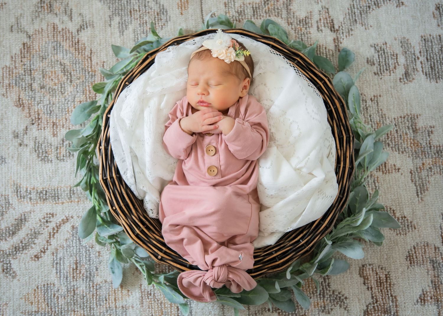 Sleeping newborn in pink swaddle rests peacefully in rustic woven basket with green leaves and white fabric.