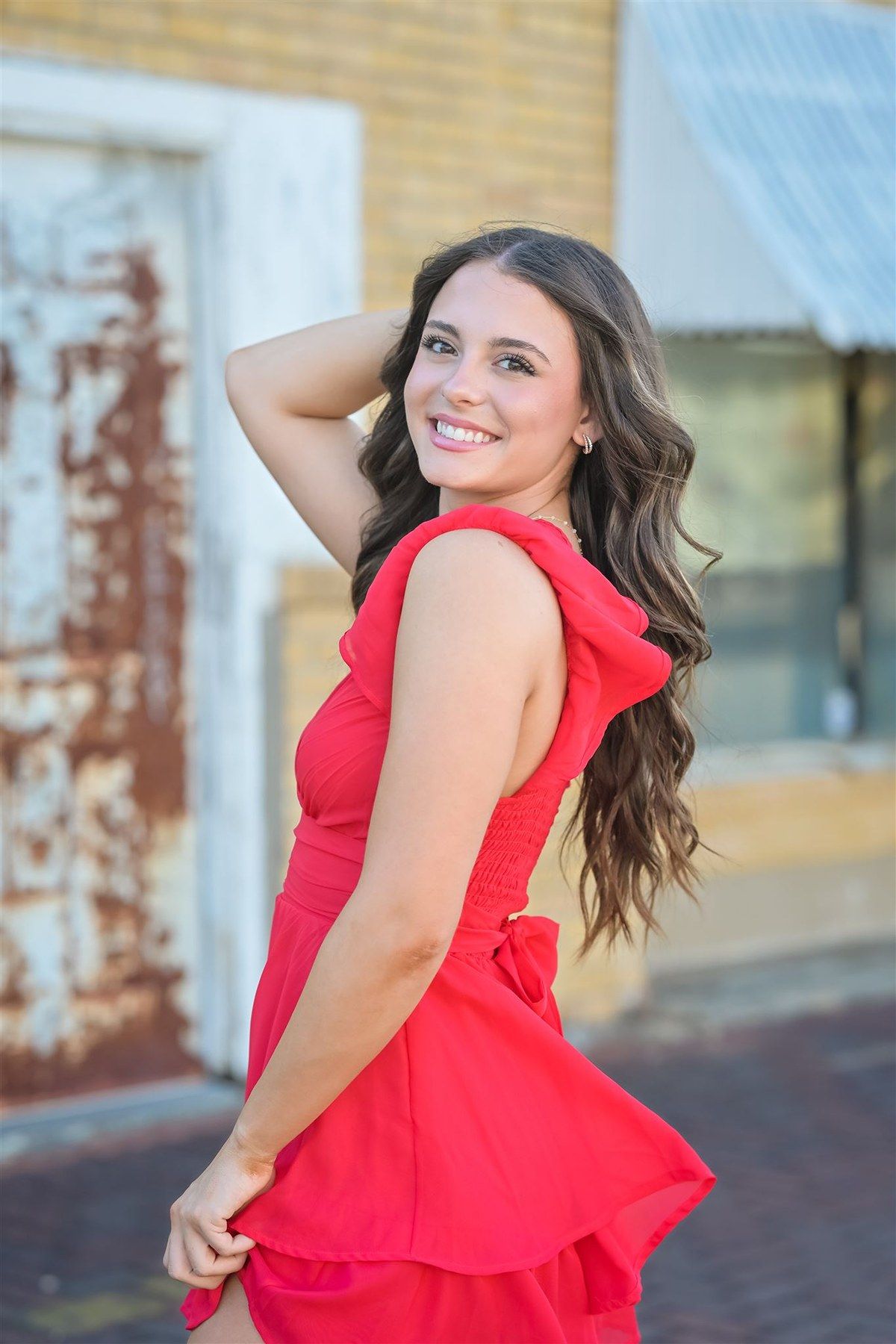 Young woman in bright red sleeveless dress poses against brick building in outdoor photo series.