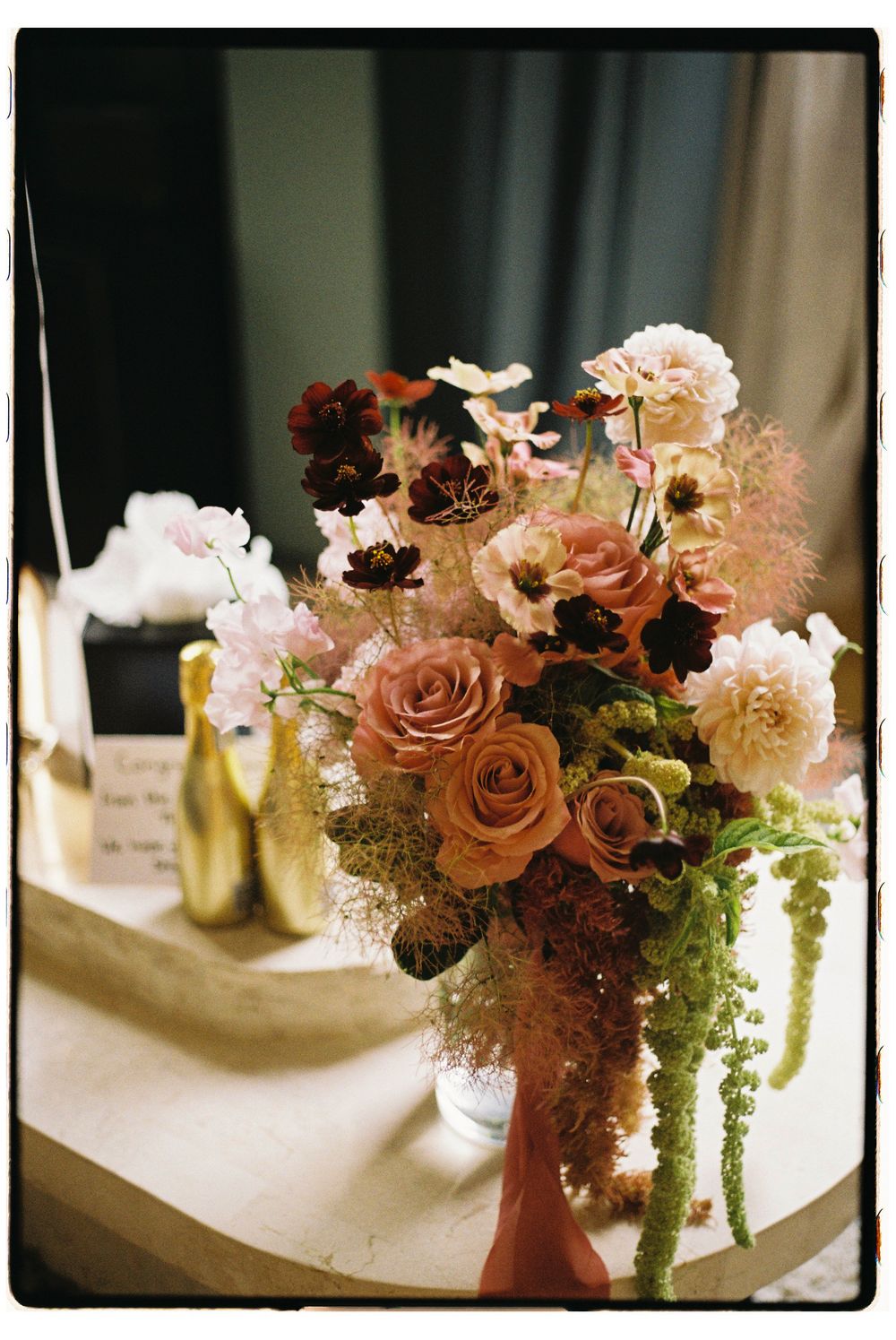 Elegant floral arrangement with peach roses and trailing greenery in a vase alongside gold wine bottles.