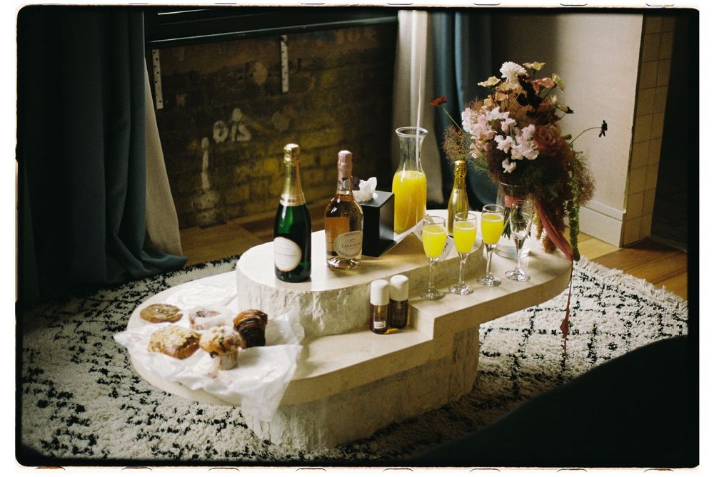 A vintage film photo of drinks and snacks arranged on a white tablecloth with a floral arrangement and fireplace in background.