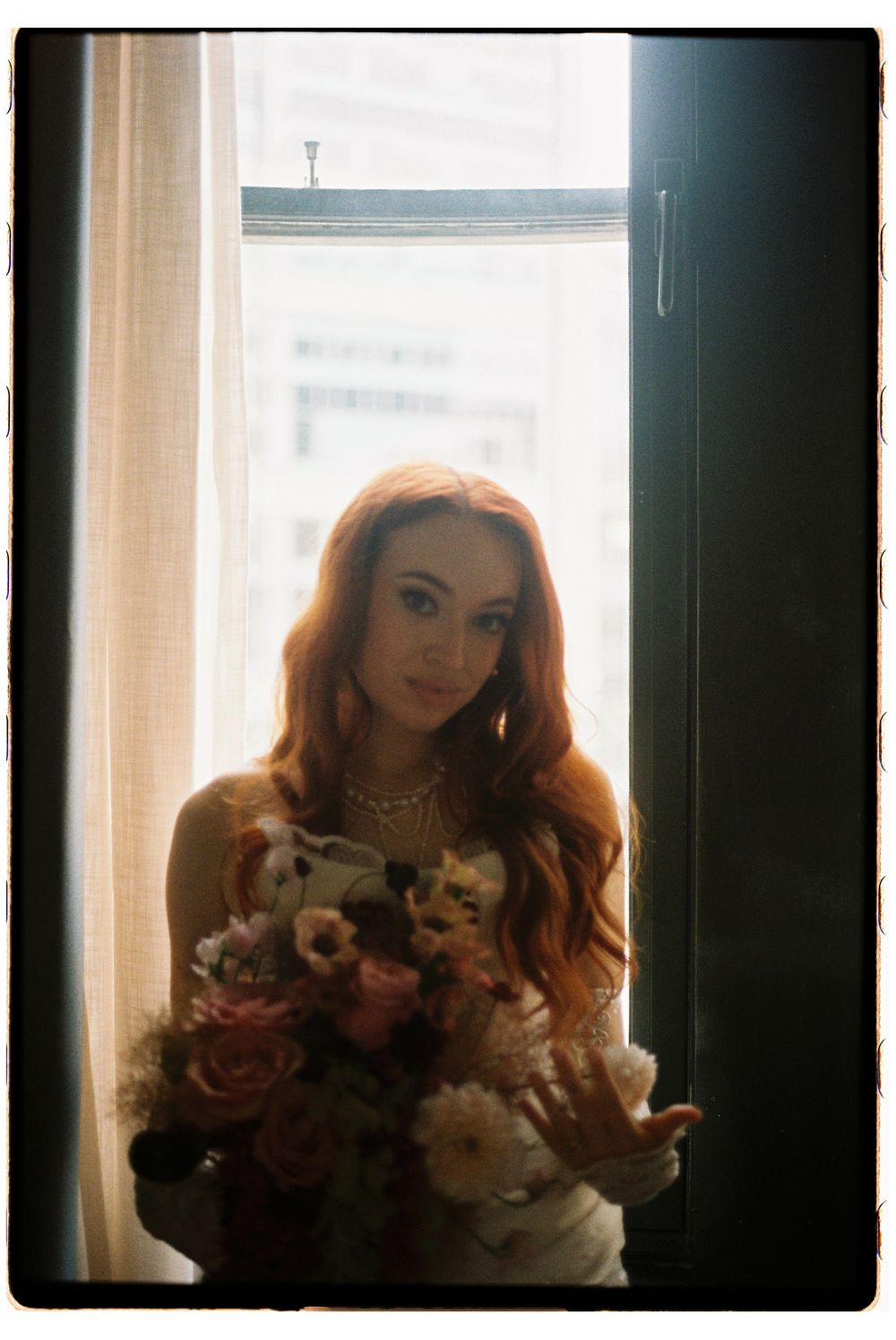 A person with long red hair stands in front of a window holding a bouquet of pink and white flowers.