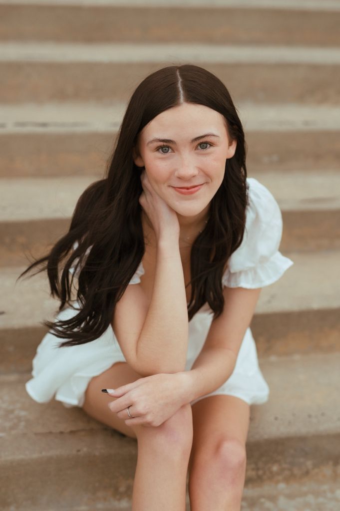 A person in a white flowy dress sits on concrete stairs, resting head on hand in a casual outdoor portrait.
