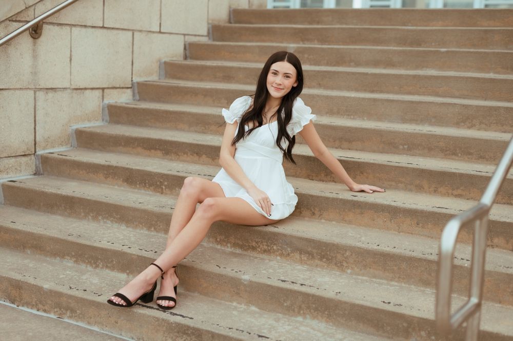 Person in a white dress and black sandals poses gracefully on concrete steps in outdoor photoshoot sequence.