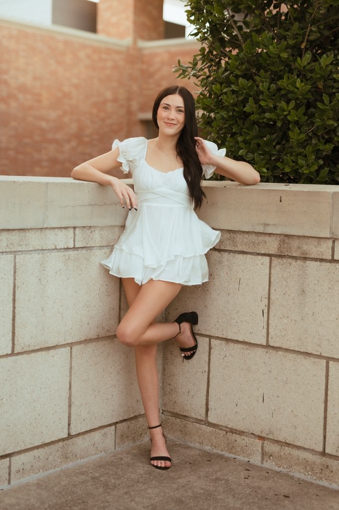 A woman in a white ruffle dress and black heels poses against a concrete wall near greenery.