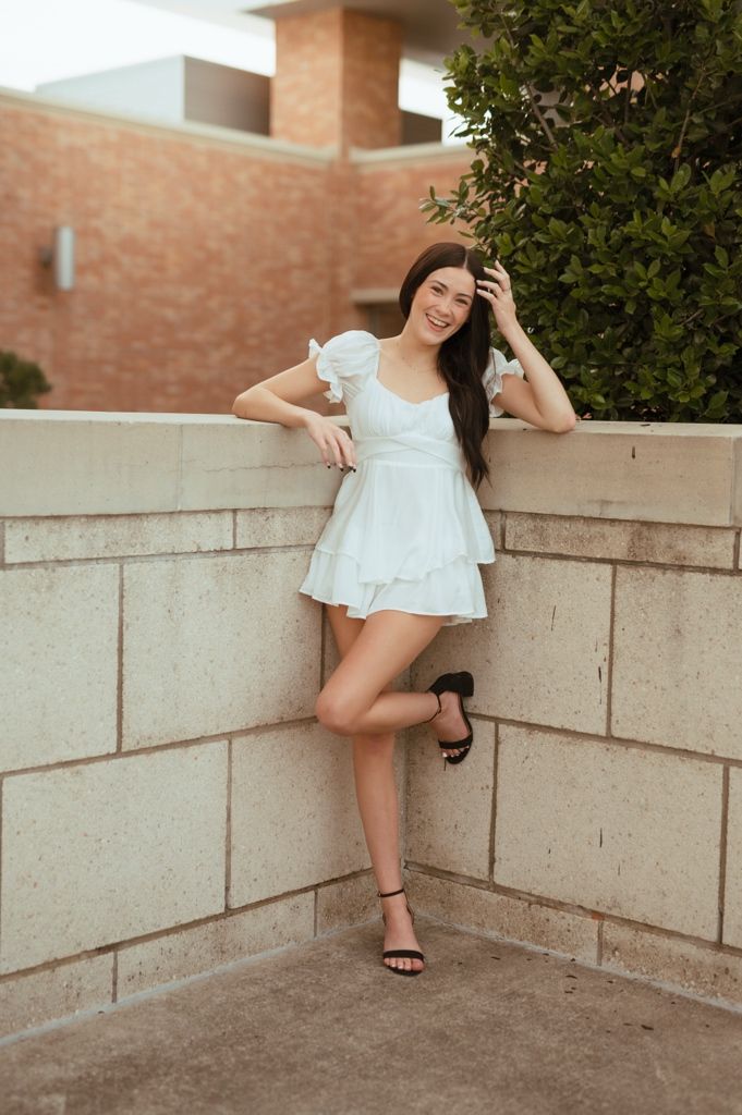 A woman in a white summer dress and sandals leans casually against a beige wall outdoors.
