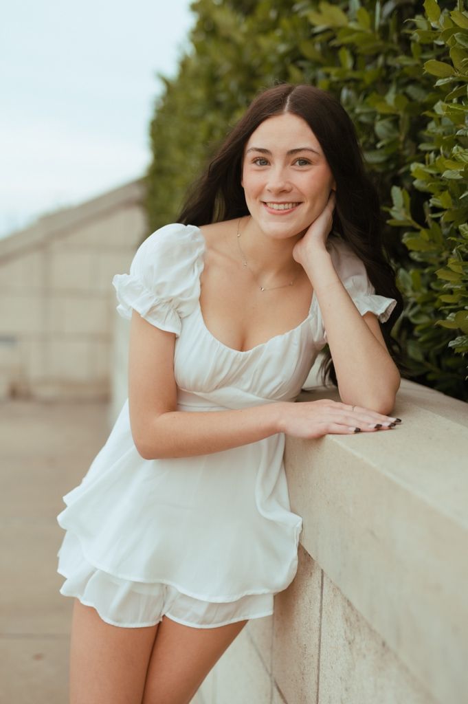 A person in a white ruffled dress leans on a light colored wall outdoors near greenery.