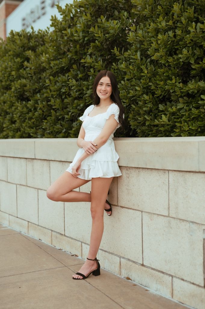 Multiple shots show a seated figure in a white ruffled top and sneakers posing against a beige block wall outdoors.