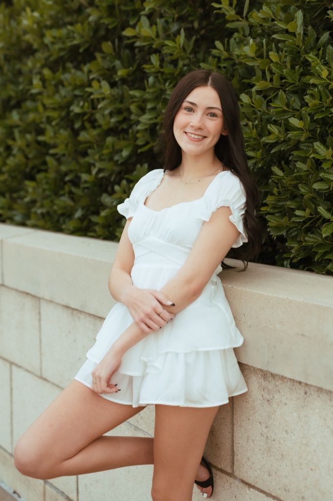 Young woman in white summer dress poses by green hedge wall on sunny day.