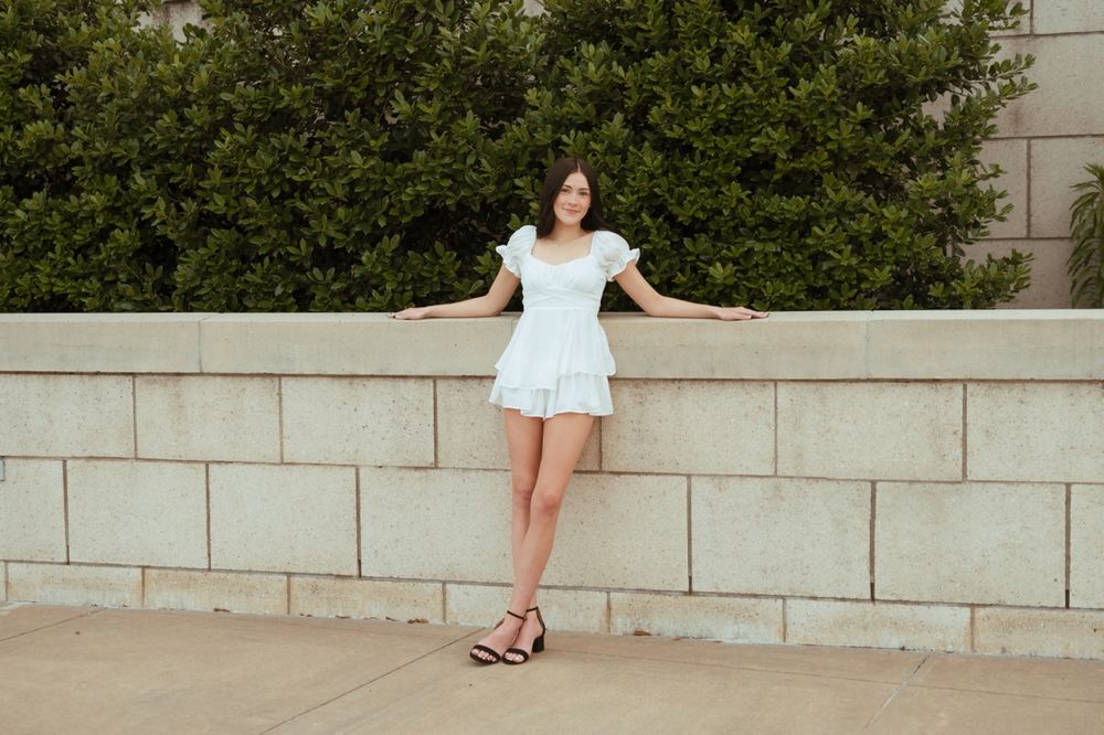 A person wearing a white flowy dress sits on stone steps surrounded by green foliage behind a retaining wall.