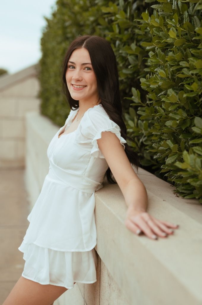 Portrait of woman in white ruffle dress leaning against wall next to green shrubs.