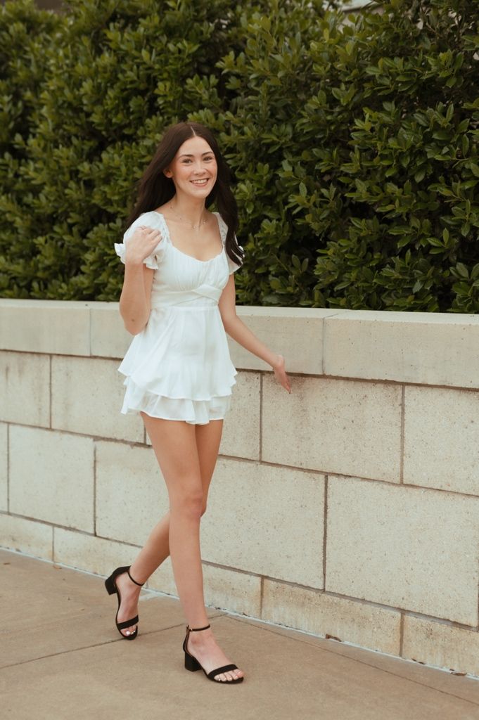 Someone wearing a white summer dress and black sandals poses against a stone wall near green bushes.