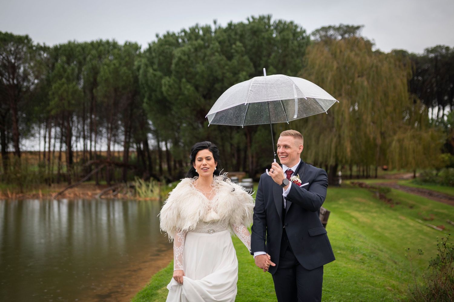 Two people laughing together while sharing a clear umbrella on a rainy day near a lake with pine trees.