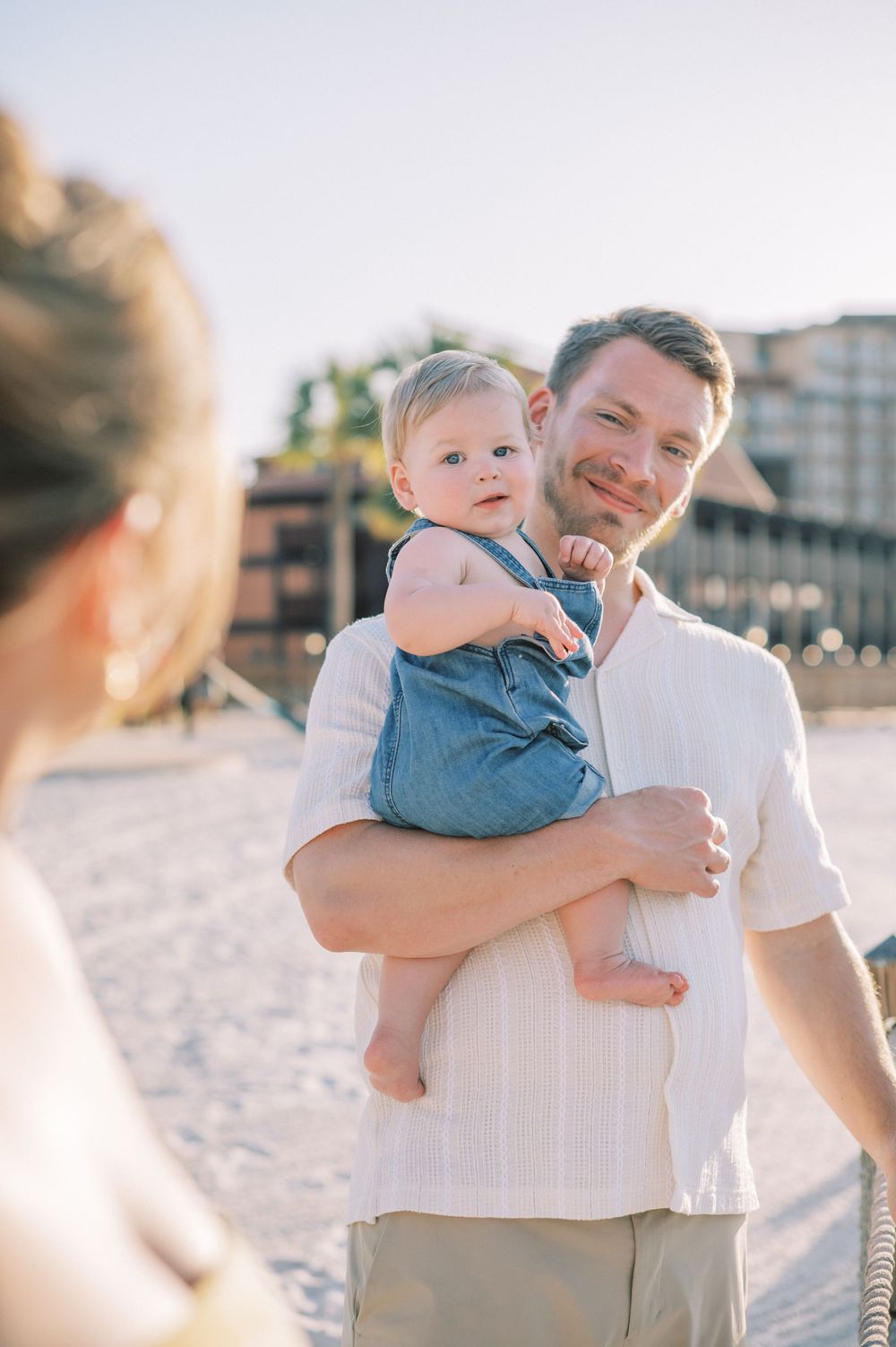 A heartwarming beach portrait shows a baby in blue denim overalls being held during golden hour photography.